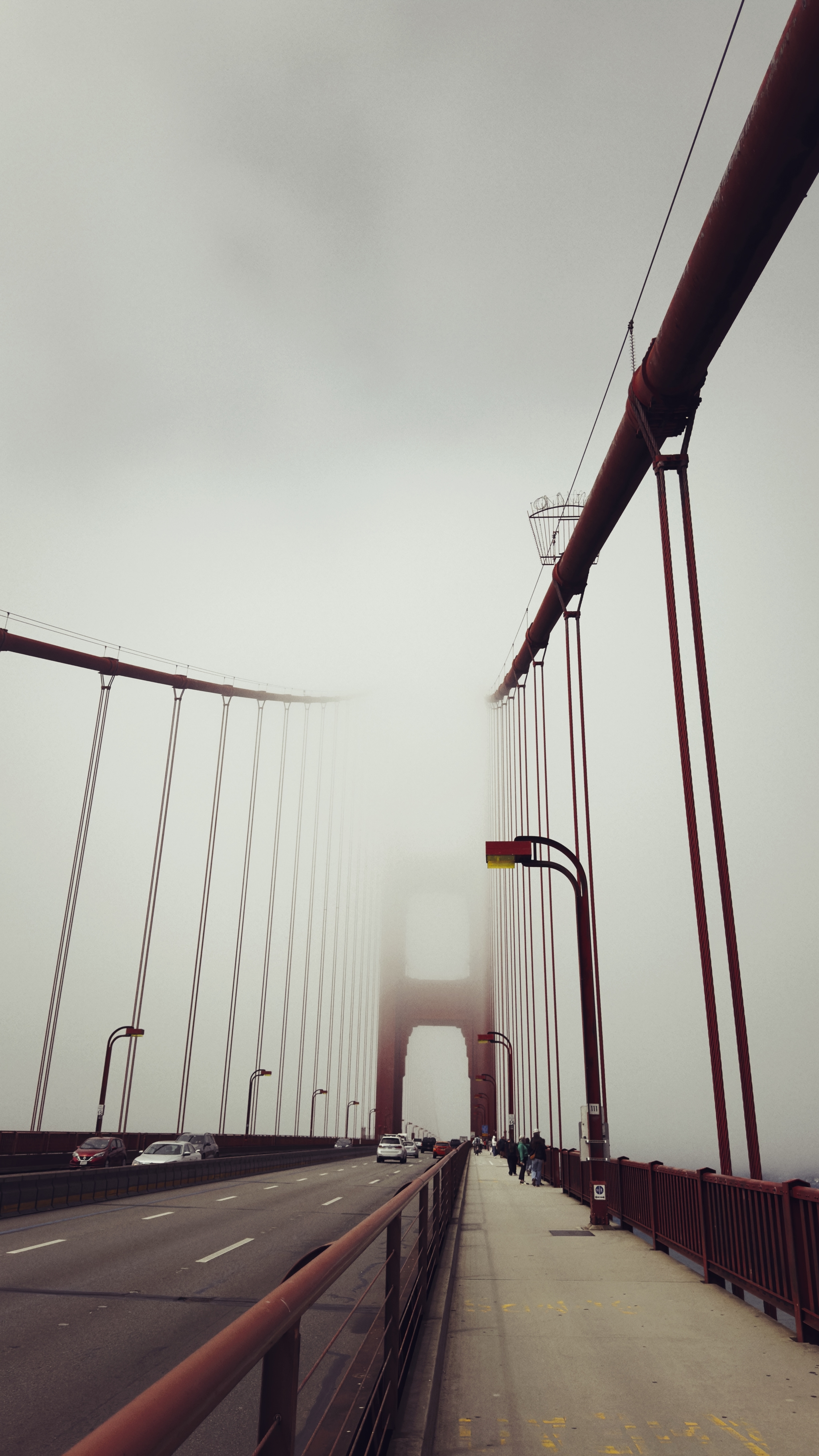 A foggy view of a suspension bridge with red cables and towers, featuring a road and pedestrian walkway