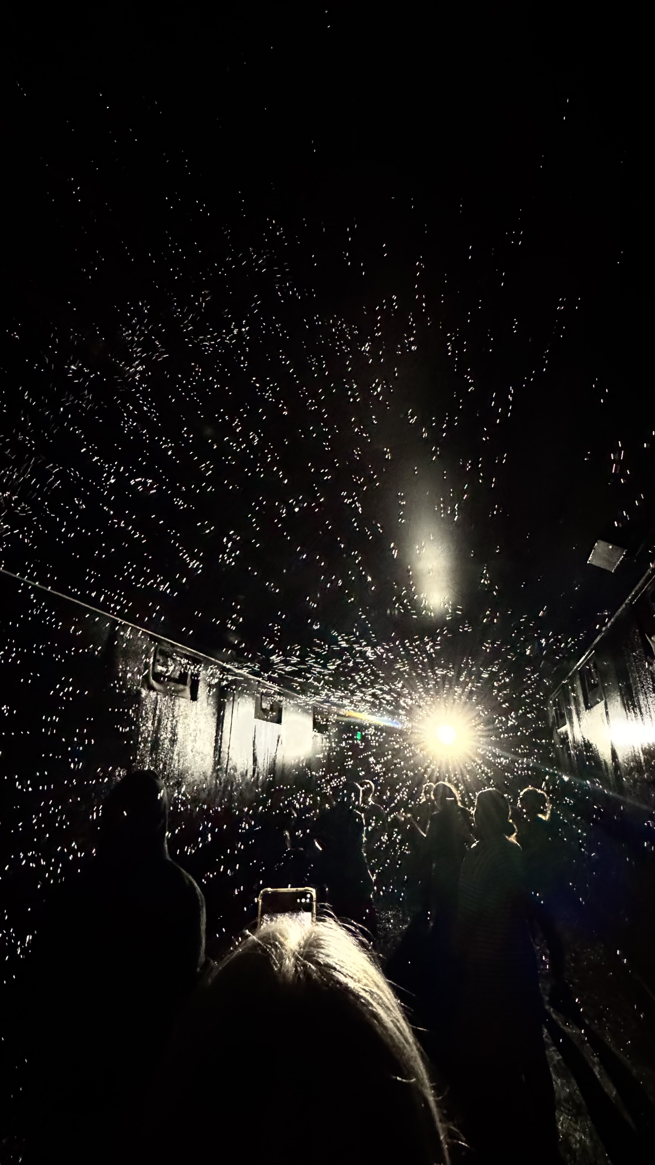 A dark tunnel with a bright light at the end, silhouettes of people, and water droplets reflecting the light, creating a starry effect