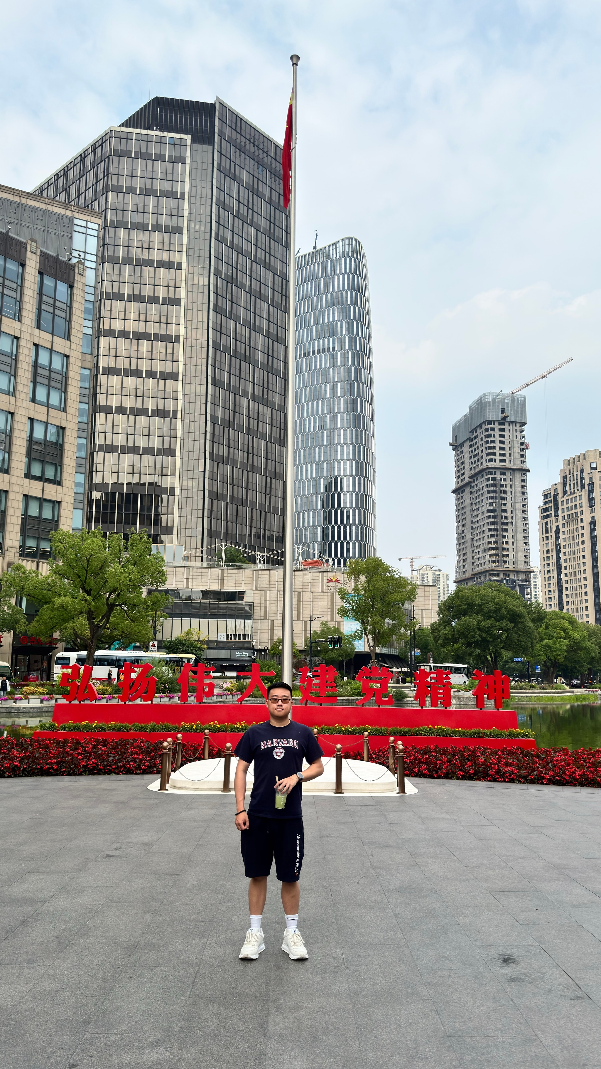 A person stands in front of a flagpole with a red flag, surrounded by modern skyscrapers and red floral arrangements
