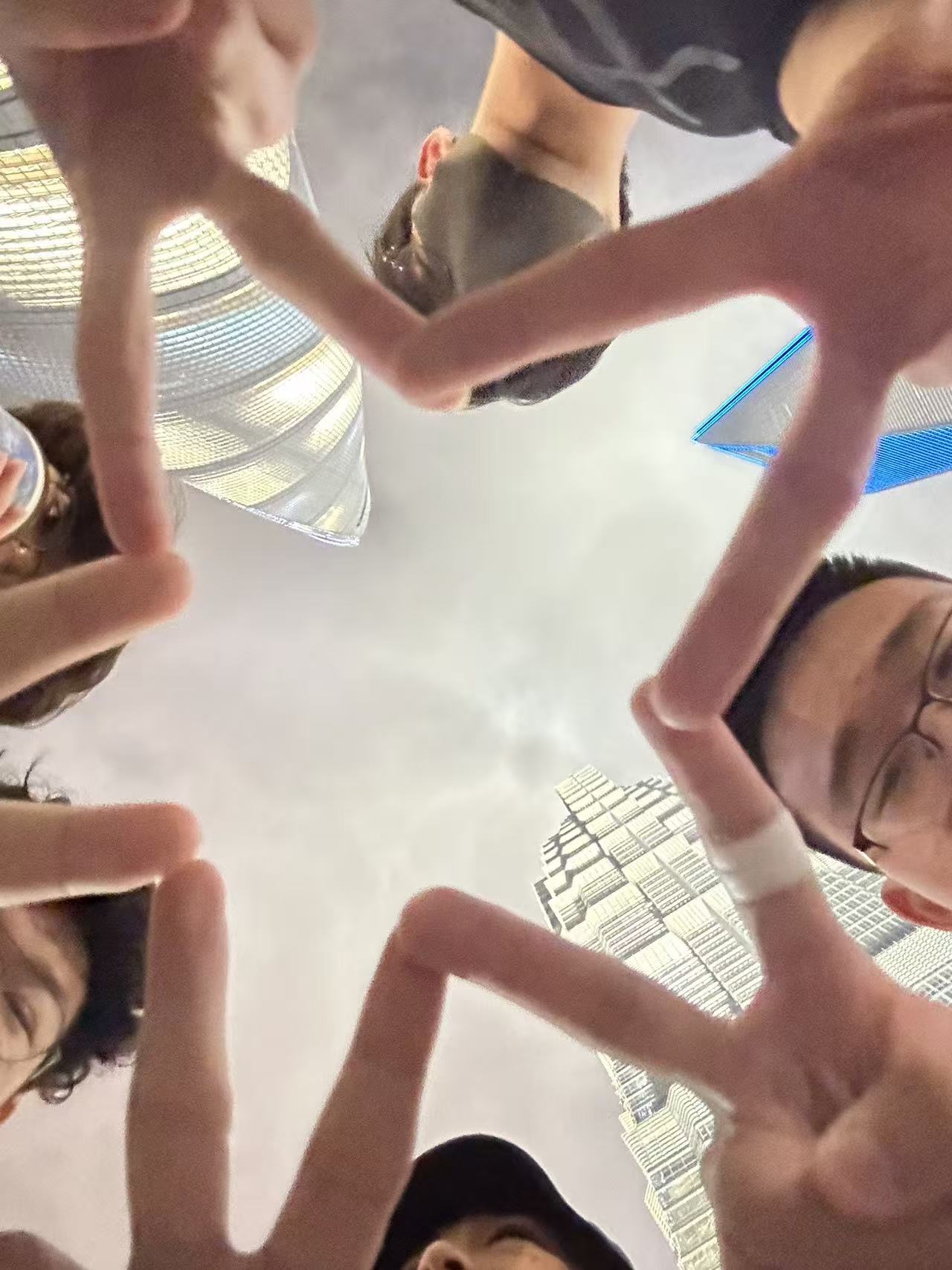 A group of people forming a star shape with their fingers, viewed from below, with tall buildings in the background