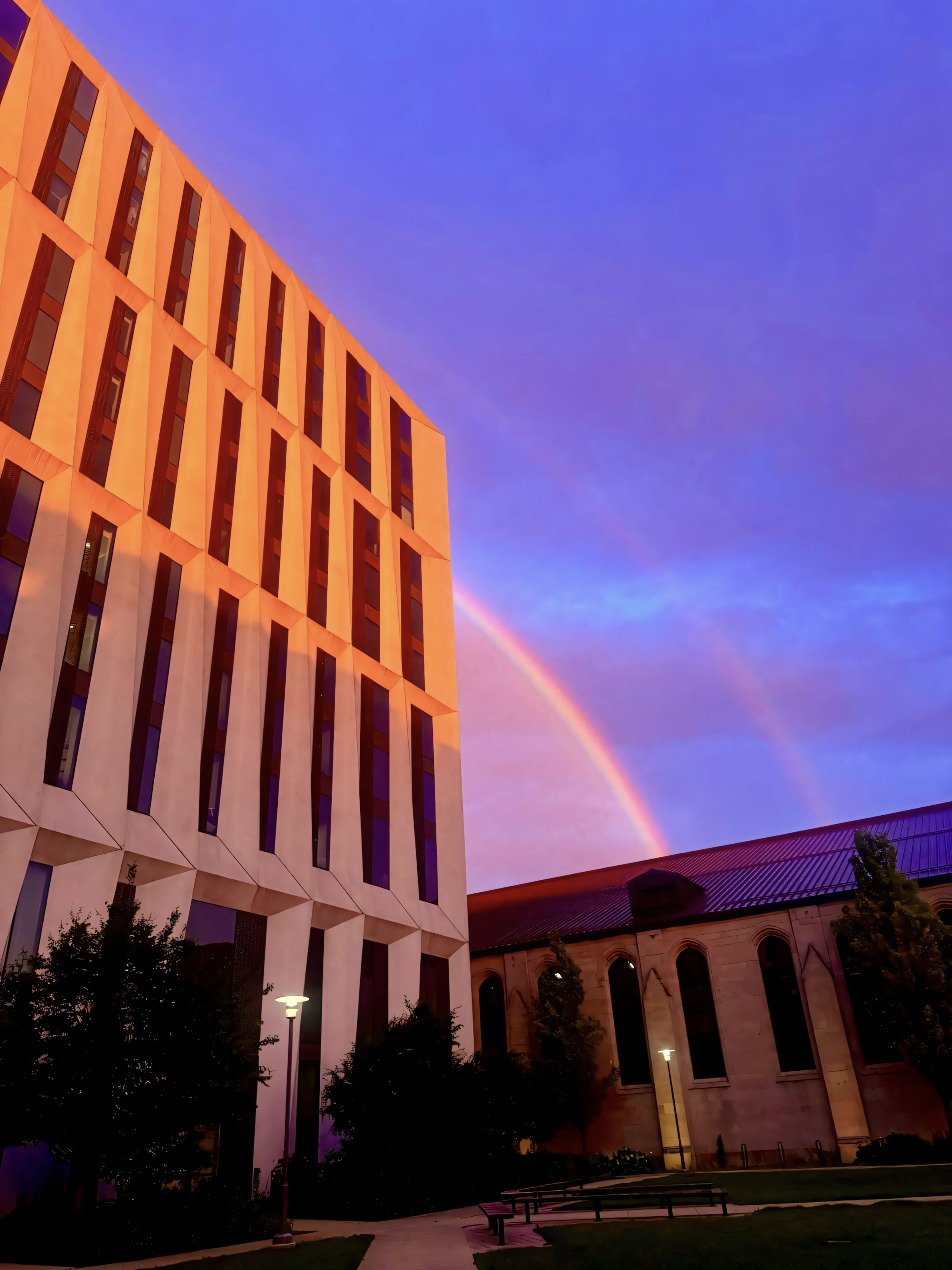 A modern building with vertical windows is illuminated by a warm light, set against a vibrant sky featuring a double rainbow