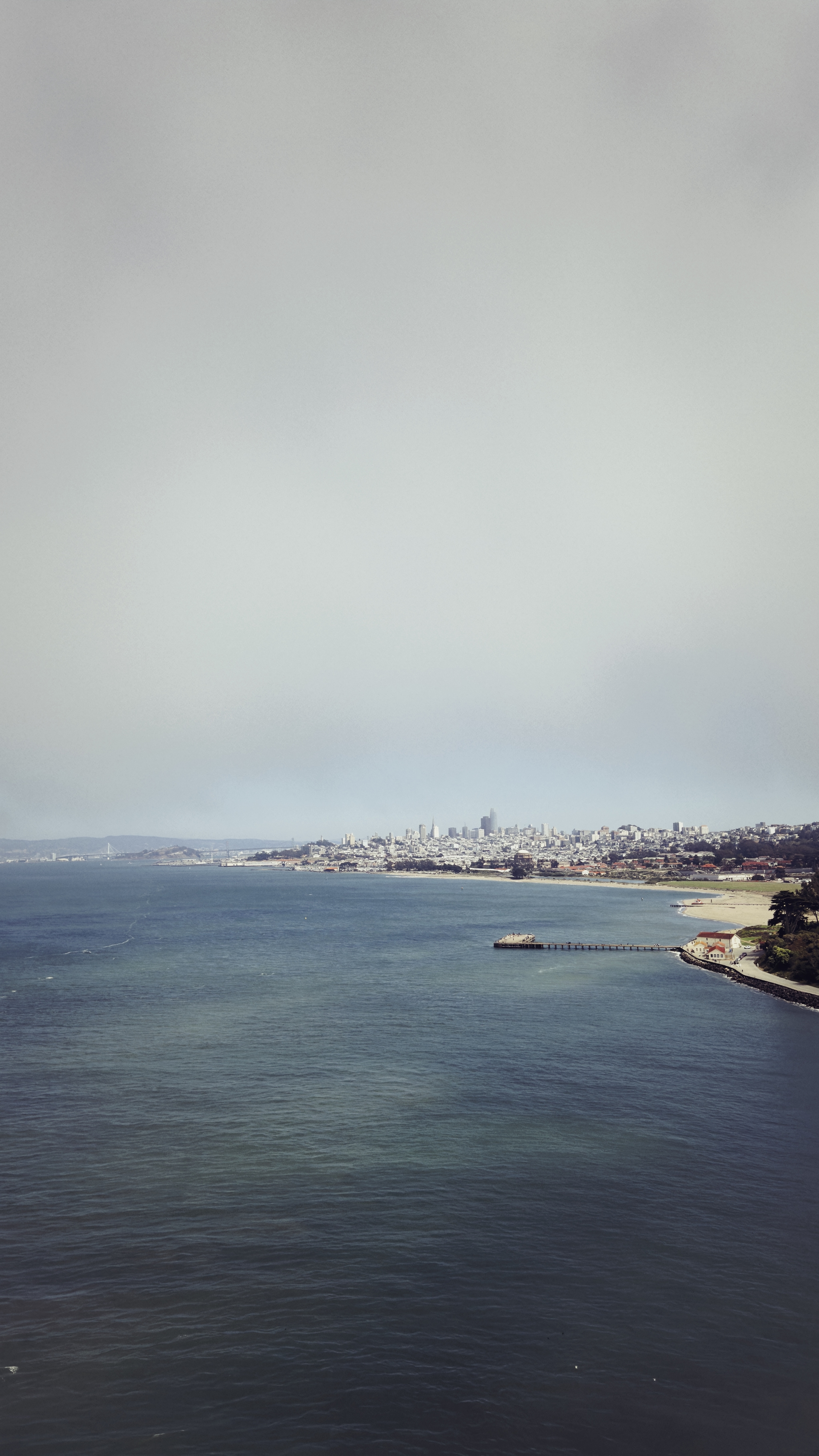 A coastal cityscape with a cloudy sky, featuring a body of water in the foreground and buildings along the shoreline in the distance