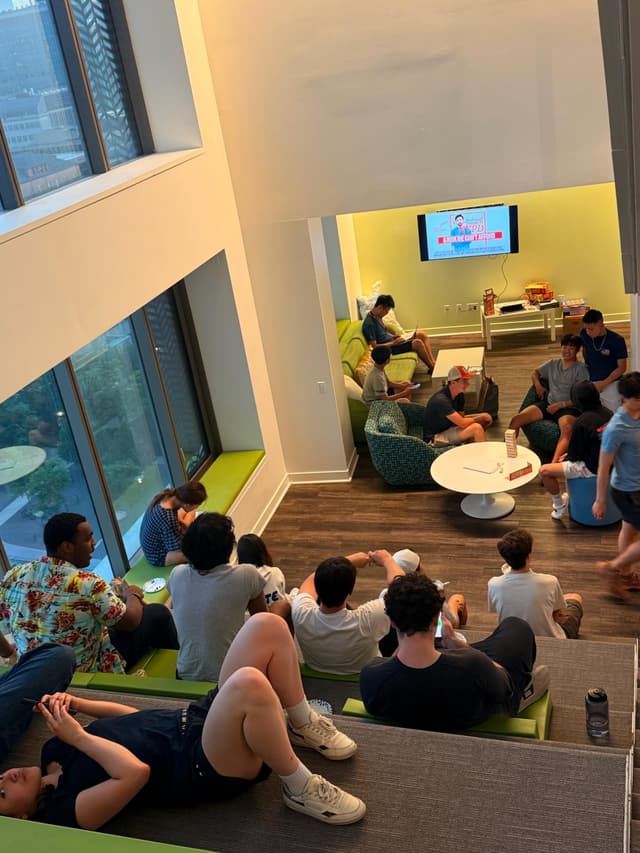 A group of people are gathered in a modern, multi-level lounge area, watching a screen Some are seated on steps, while others are on couches and chairs The room has large windows and a bright, open atmosphere