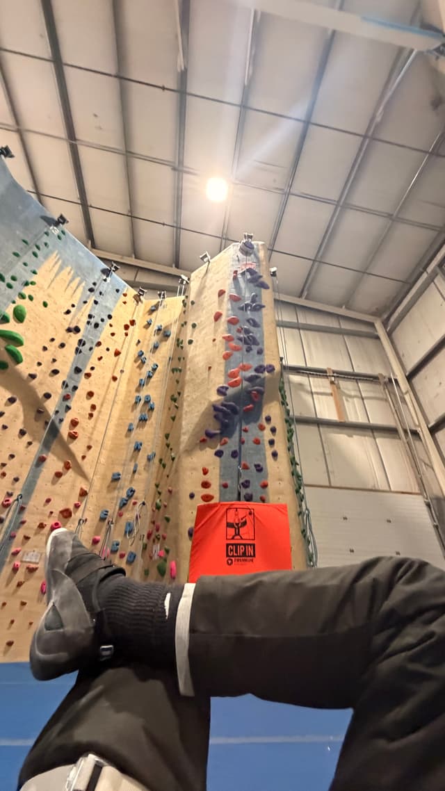 A person is sitting with legs crossed, wearing climbing shoes, in front of an indoor climbing wall with colorful holds and a crash pad at the base
