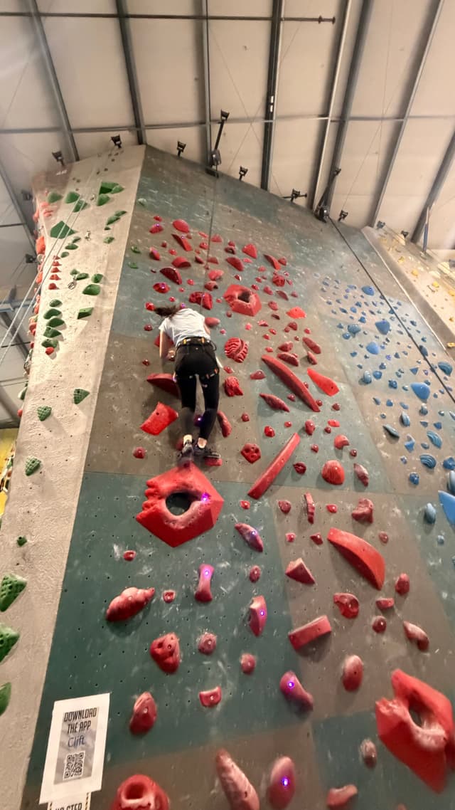 A person climbing an indoor rock wall with various colored holds, including red, green, and blue, under a high ceiling