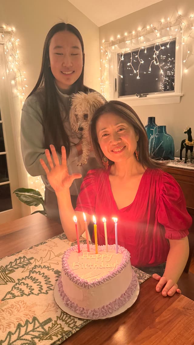 A woman in a red dress is sitting at a table with a lit birthday cake, smiling and waving. Another person stands behind her holding a small dog. The room is decorated with string lights