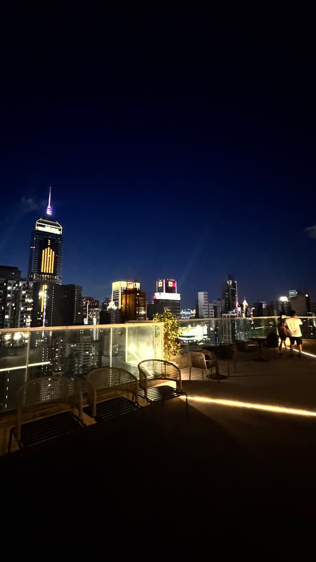 A city skyline at night with illuminated skyscrapers and a rooftop terrace featuring seating and ambient lighting