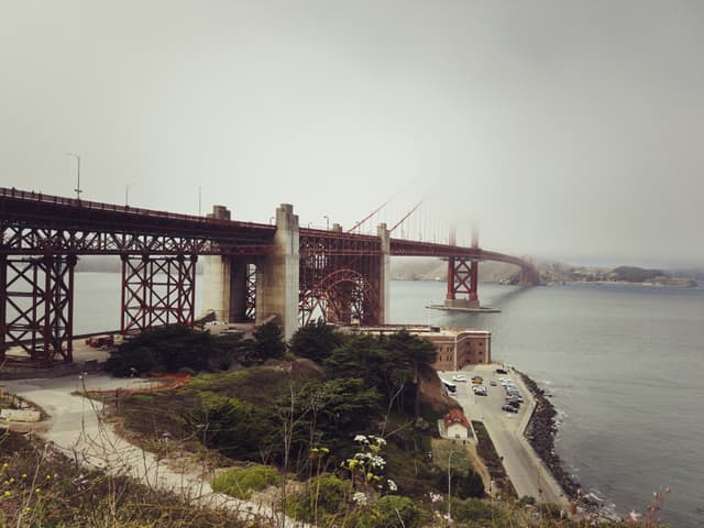A large suspension bridge spans across a body of water, with a foggy sky and a view of the shoreline below
