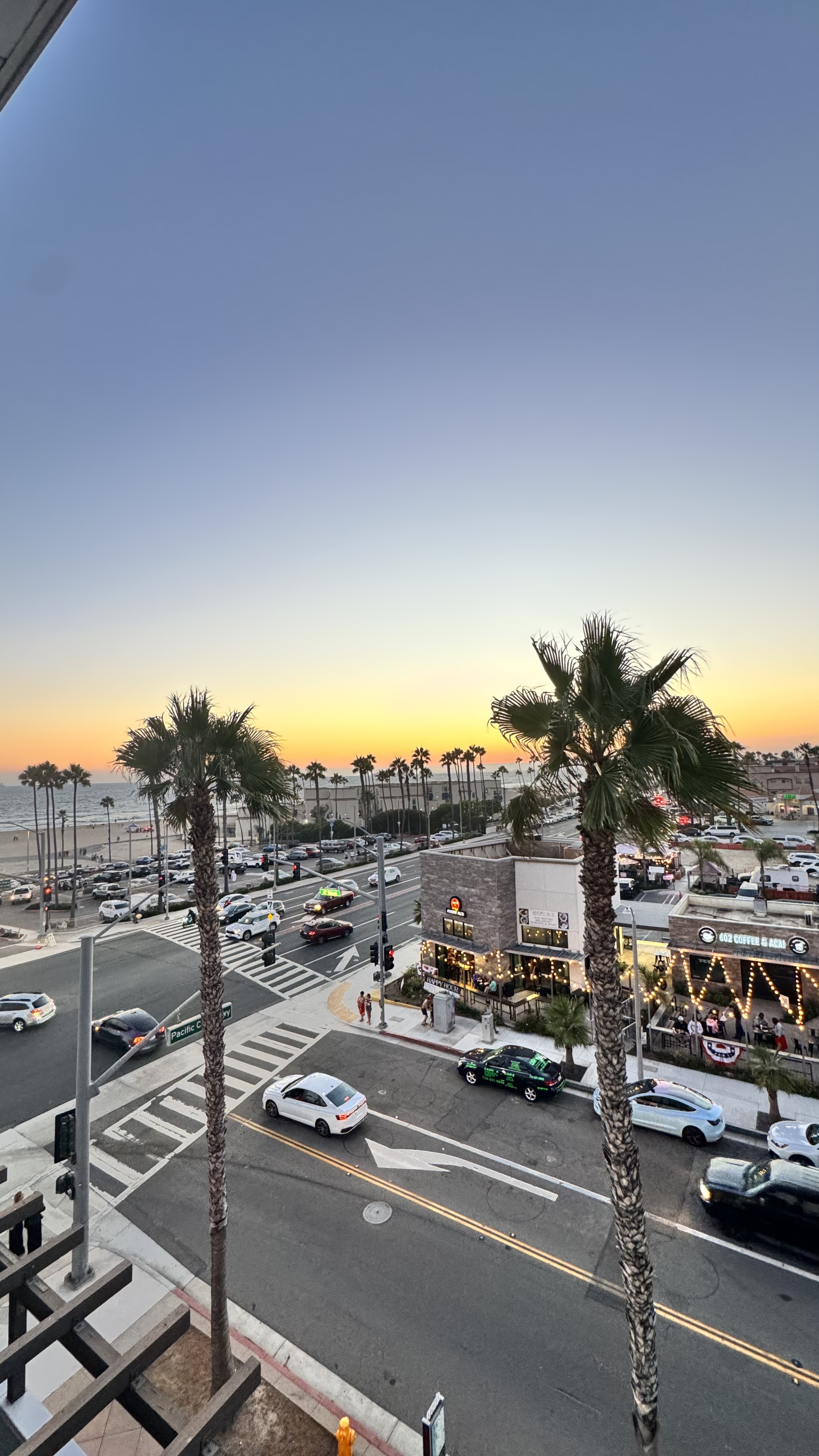 A coastal street scene at sunset with palm trees, cars, and a view of the ocean in the background